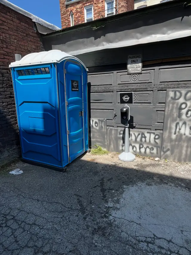 One blue standard porta potty set beside a building wall on asphalt