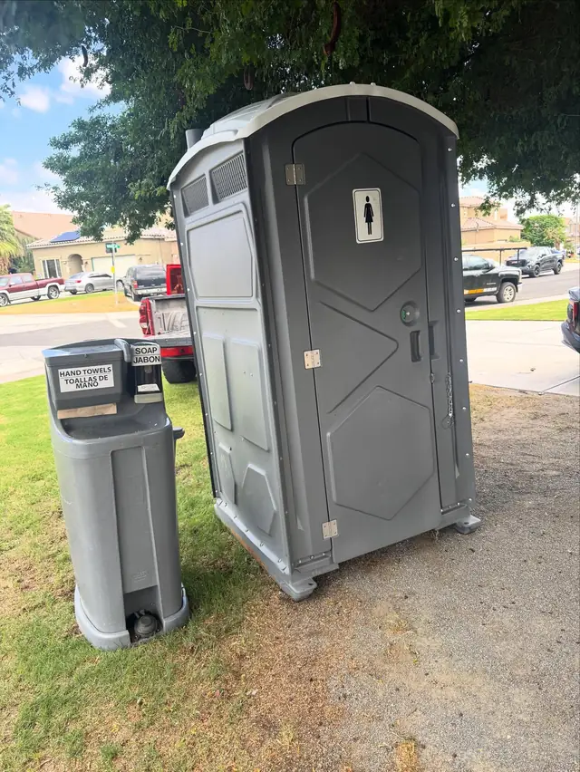A gray standard porta potty and a hand wash station set level on a backyard lawn